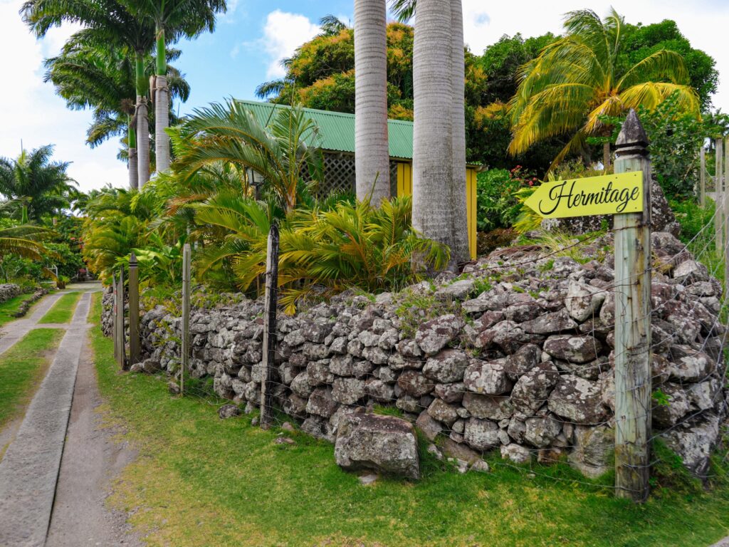 A painted sign on a timber post beside a dry-stone wall, with a narrow lane running past palms towards a green-roofed cottage.