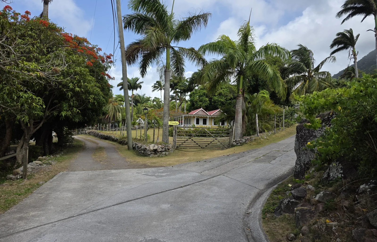 A yellow Hermitage sign by a wooden gate and dry-stone wall, with a pink-roofed cottage among tall palms beside a concrete lane.