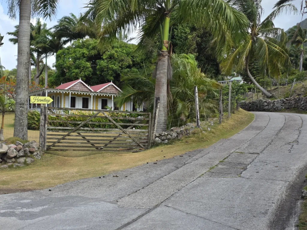 A concrete road curving away to the right past tall palms, with a red-roofed gingerbread cottage behind the gate.