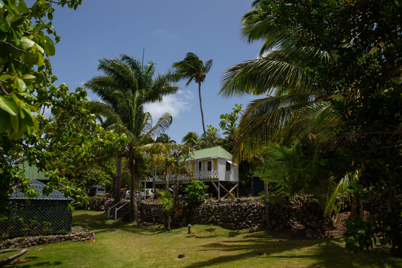 A green-roofed cottage raised on posts in the estate garden, with palms and the hillside rising behind it.