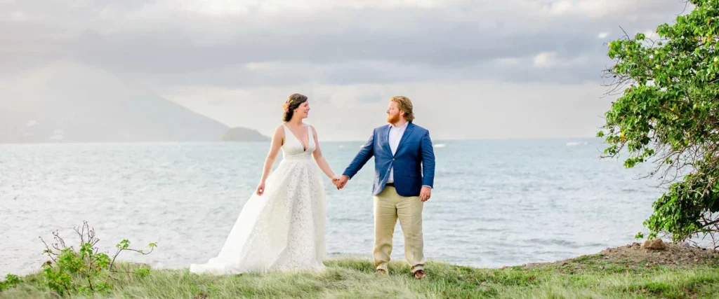 A newly married couple in wedding attire hold hands on a grassy shoreline in Nevis, with the sea and hills behind them