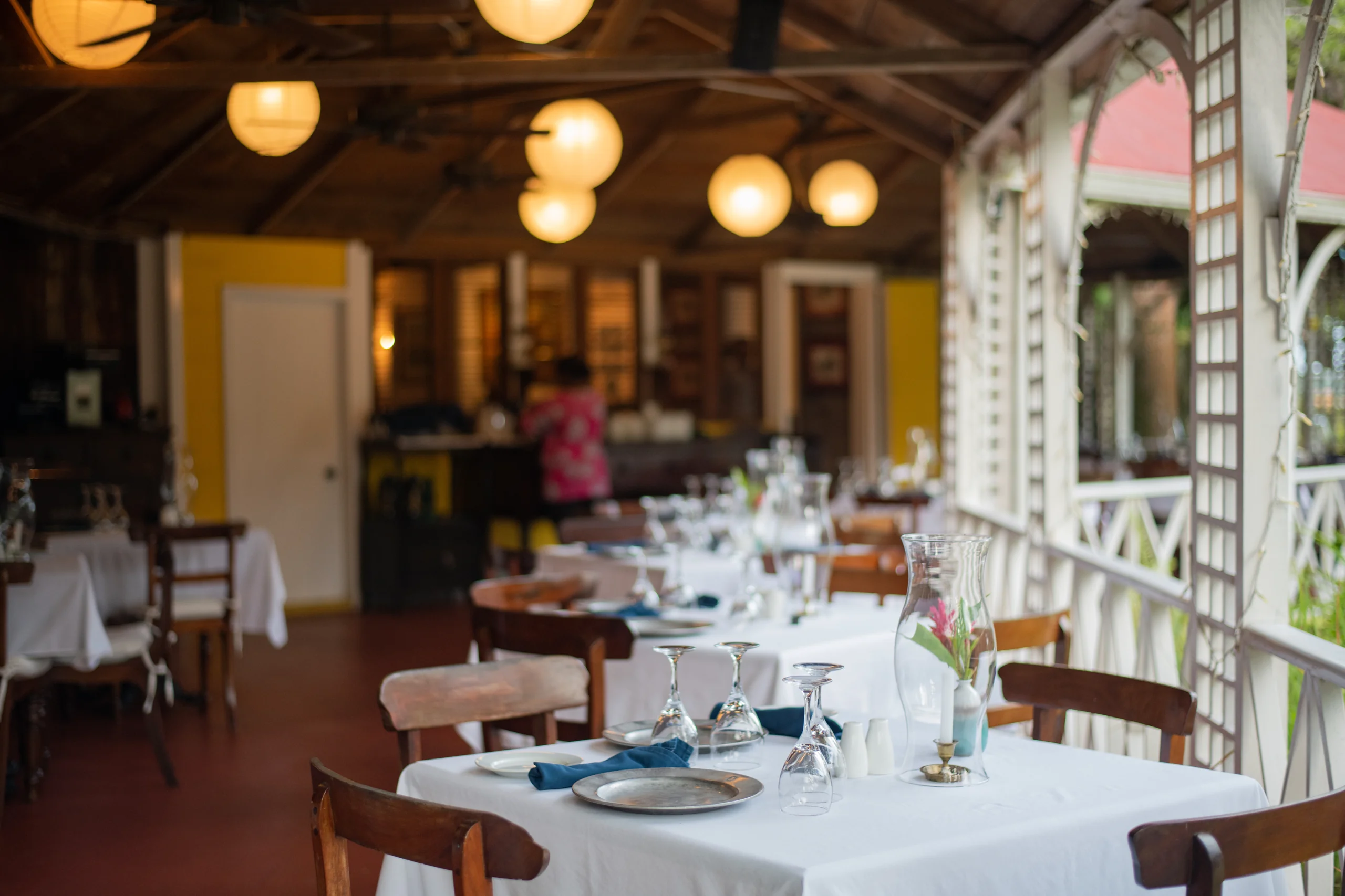 Tables laid with white linen on the dining verandah at Hermitage Nevis, beneath hanging paper lanterns and the open timber roof
