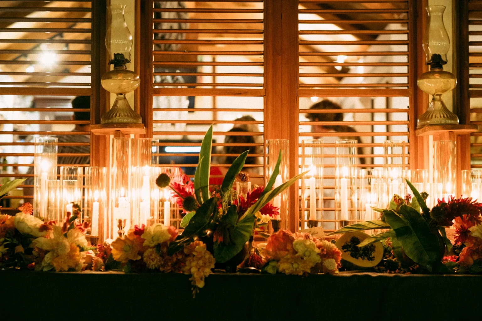 Candles in tall glass hurricanes and tropical flowers with anthurium leaves along a celebration table, with oil lamps and wooden shutters behind
