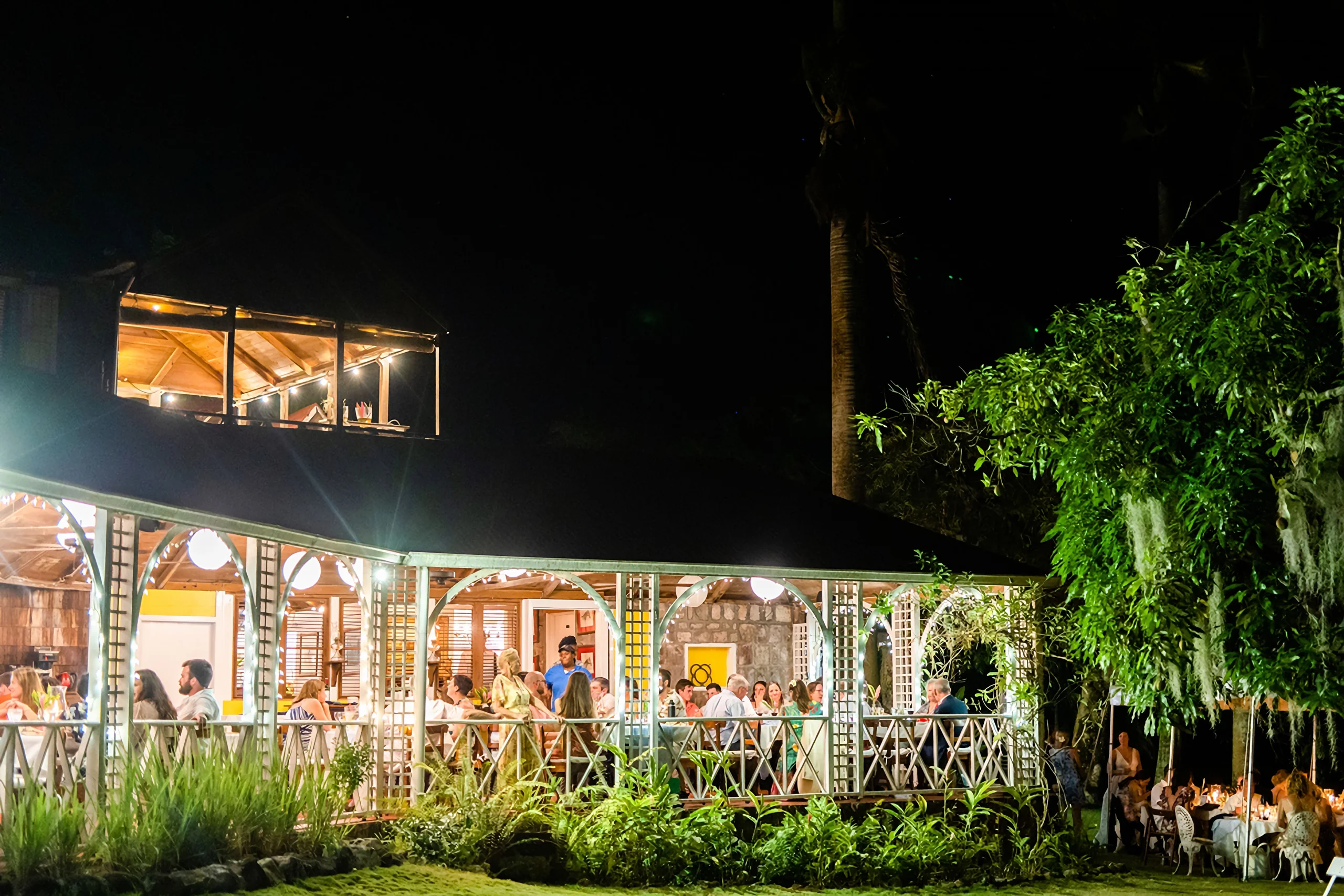 Evening at Hermitage Nevis: guests gathered at long tables on the lit gingerbread verandah of the Great House, with more tables set out under the trees on the lawn