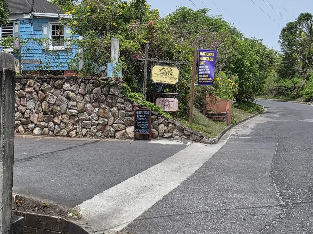 View from the main road of the turning onto Hermitage Road, beside a dry-stone wall and a blue shingled house, with the hotel signs on the corner.